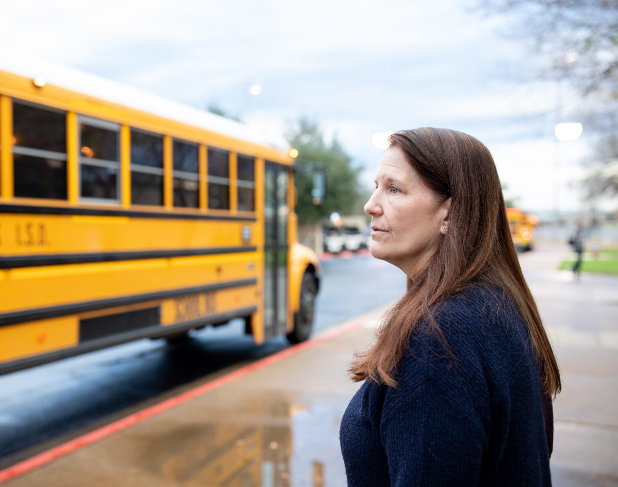 A woman with long brown hair, wearing a dark sweater, stands outside looking thoughtfully at a yellow school bus as it drives away. The bus is labeled 'Round Rock I.S.D.' and the scene takes place on a cloudy day with damp pavement.